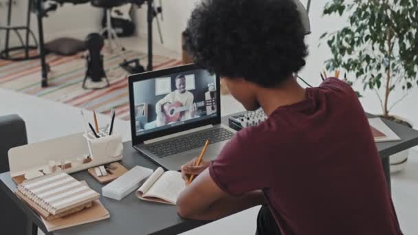 Задний вид кудрявого молодого человека, сидящего за столом в своей комнате утром, делающего заметки во время занятий онлайн-музыкой на портативном компьютере с черным человеком, который играет на гитаре - Кадры, видео