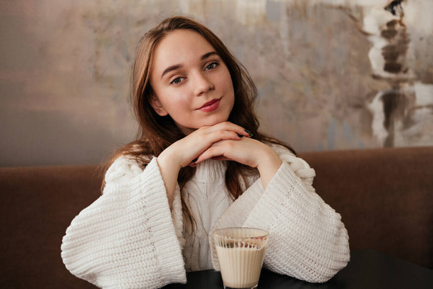 Jovencita sonriente sentada en la cafetería y tomando café. Caucásico chica en suéter en el interior - Foto, imagen