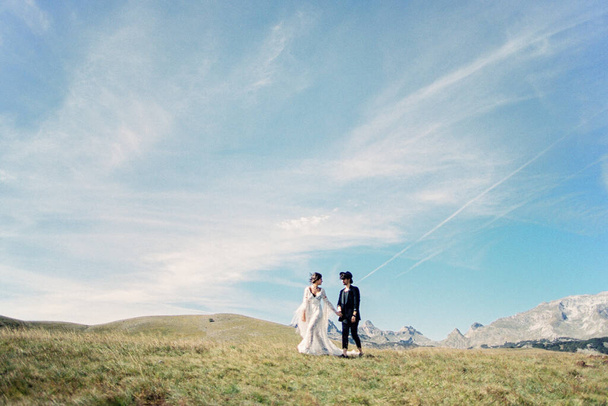 A menyasszony és a vőlegény kéz a kézben sétálnak a zöld pázsiton a hegyek hátterében. - Fotó, kép