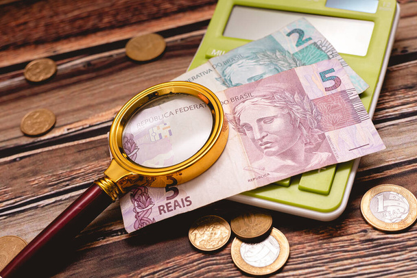 A golden magnifying glass over Brazilian Real banknotes. Coins and a blue calculator in the image composition. - Foto, Imagen