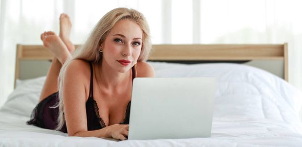 zbliżenie studio strzał z biały kobieta blondynka włosy Gorąca kobieta moda model w czarny sexy bielizna bielizna leżał w dół uśmiechnięty przeglądanie surfing internet online przez laptop na łóżko w sypialni - Zdjęcie, obraz