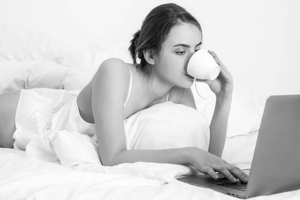 Fiatal nő jól érzi magát az ágyban a nappaliban, kávé és laptop. - Fotó, kép