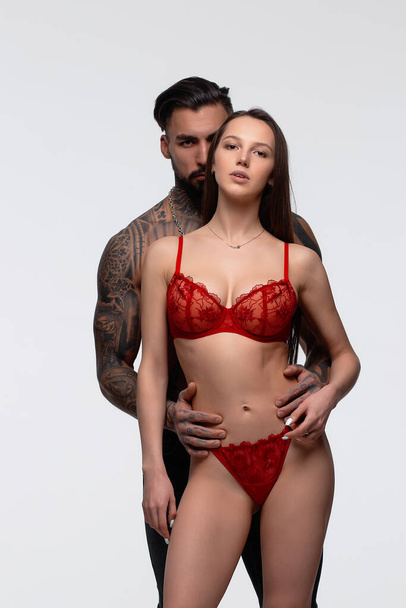 Fiducioso maschio barbuto tatuato in piedi con seducente partner femminile che indossa lingerie di pizzo rosso in studio e guardando la fotocamera - Foto, immagini