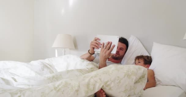 Echemos un vistazo antes de levantarnos. 4k video filmación de un joven padre guapo y su hijo usando una tableta en la cama en casa. - Metraje, vídeo