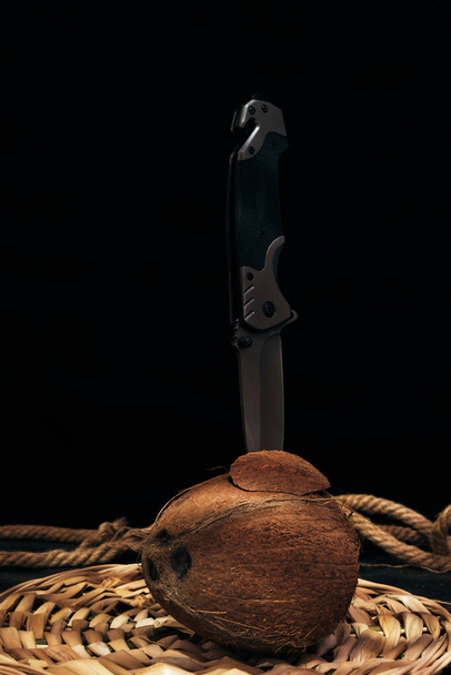 Tactical folding knife is stuck in split coconut lying next to pano woven from seeds.Survival and adventure - Photo, Image