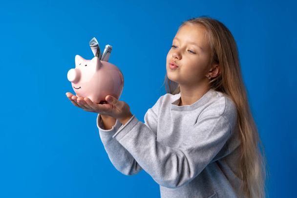 Beautiful little girl with piggy bank on blue background - 写真・画像