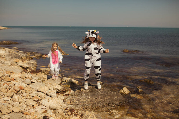 Two pretty girls in kigurumi cow and unicorn walk on the seashore, friends chat and play - Photo, Image