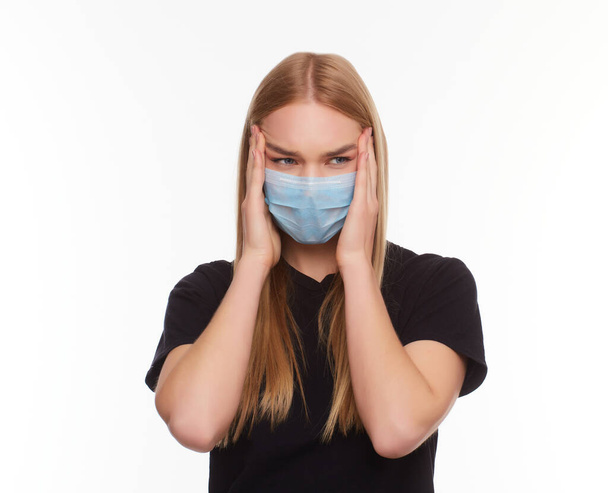 Kapalı gözlü, tıbbi grip maskesi takan, başını eliyle tutarak beyaz arka planda izole edilmiş genç sarışın kızın stüdyo portresi.. - Fotoğraf, Görsel