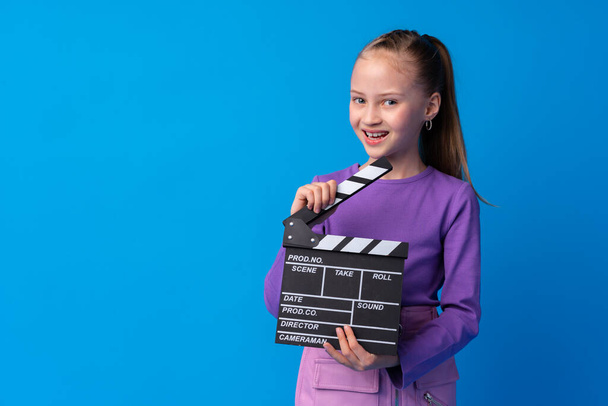 Smiling girl holding clapper board against blue background - Фото, изображение