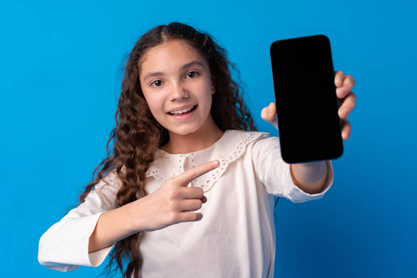Young girl with dark curly hair showing smartphone screen against blue background - Valokuva, kuva