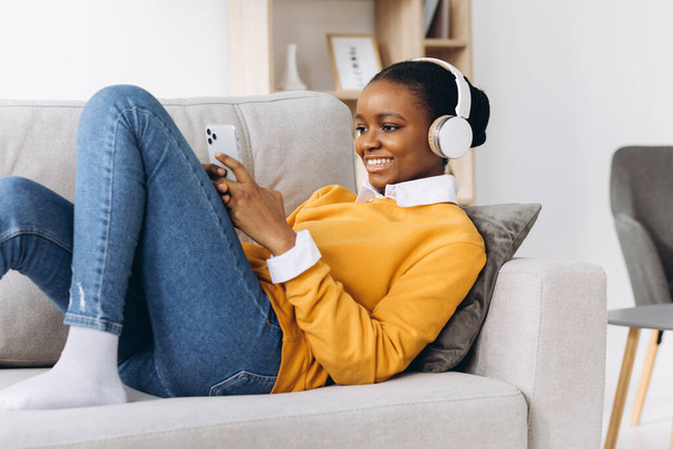 Menschen, Technik und Freizeitkonzept - fröhliche junge Afroamerikanerin sitzt mit Smartphone und Kopfhörer auf dem Sofa und hört zu Hause Musik - Foto, Bild