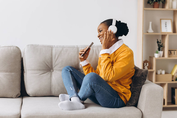 ihmiset, teknologia ja vapaa-ajan konsepti onnellinen afrikkalainen amerikkalainen nuori nainen istuu sohvalla älypuhelimen ja kuulokkeiden kuunnellen musiikkia kotona
 - Valokuva, kuva