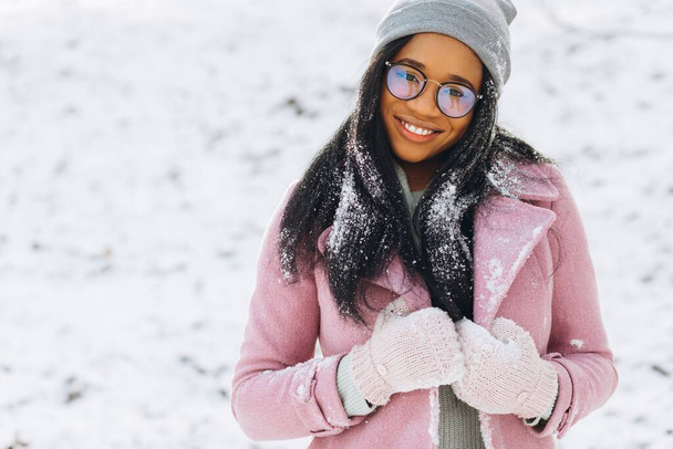 幸せな肯定的な女の子の肖像画、彼女の髪に雪と手袋でアフリカ系アメリカ人の若い女性は暖かい服で寒い霜の日に雪の中の冬の雪の公園で笑っています。冬のコンセプト - 写真・画像