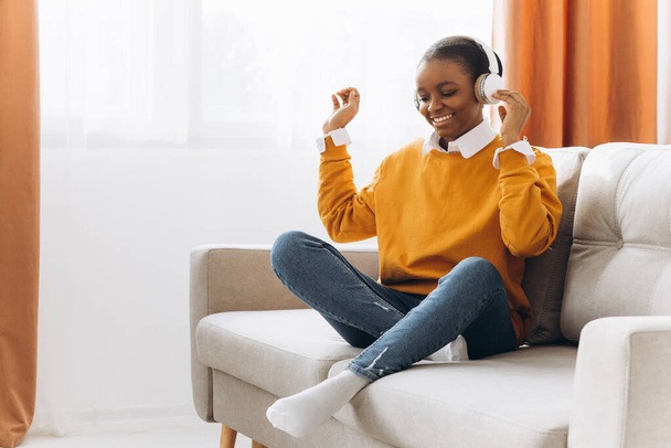 Plná délka africká žena ve sluchátkách potřást hlavou zvednout ruce a vychutnat playlist oblíbené píseň tanec v obývacím pokoji - Fotografie, Obrázek