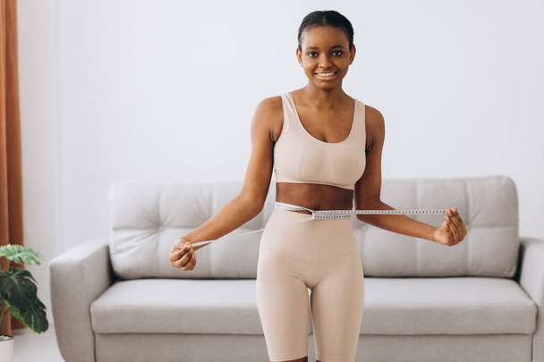 若い黒人女性はリビングルームの背景で腰を測定します。ダイエットと減量の概念. - 写真・画像
