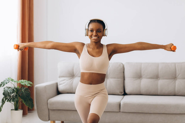 Fiatal fekete nő fülhallgatóban zenét hallgat és súlyzókkal edz otthon a nappaliban. Otthoni képzés. - Fotó, kép