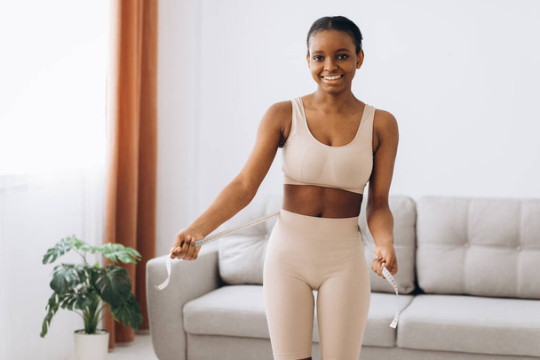 Young black woman measures her waist on the living room background. The concept of diet and weight loss. - Photo, Image