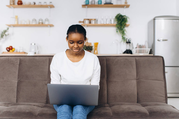 Young African American woman working on laptop sitting on sofa in kitchen - Φωτογραφία, εικόνα