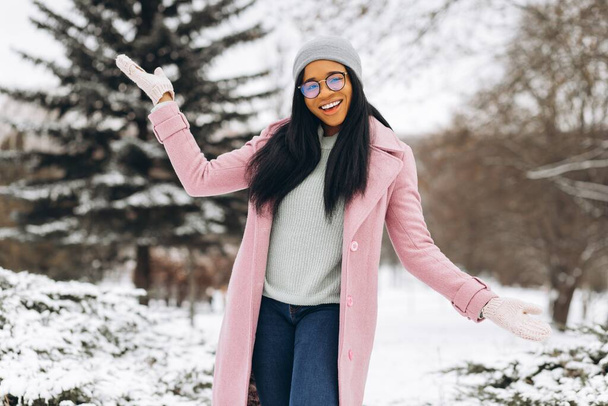 Portret van gelukkig positief meisje, Afrikaanse etnische Afro-Amerikaanse jonge vrouw in bril en handschoenen lacht in de winter besneeuwd park in de sneeuw op koude ijzige dag in warme kleren. Winter concept - Foto, afbeelding
