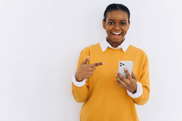 Cellulare di moda. Donna afroamericana sorridente che tiene l'ultimo smartphone sottile con schermo bianco e lo indica. - Foto, immagini