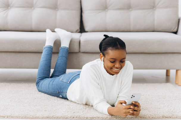 Uśmiechnięta czarna dziewczyna wiadomości na smartfonie w domu, relaks na wygodnej kanapie, korzystających weekend, przestrzeń kopiowania - Zdjęcie, obraz