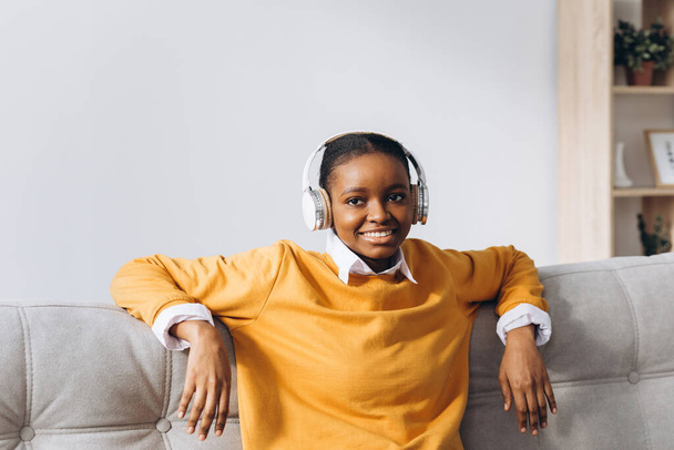 Afrikanerin in voller Länge mit Kopfhörer schüttelt erhobene Hände und genießt Playlist Lieblingstanz im Wohnzimmer - Foto, Bild
