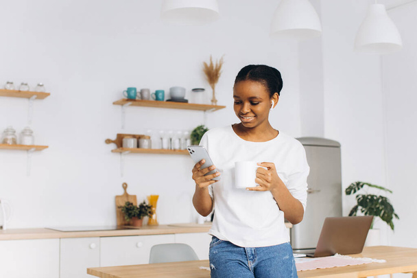 Portret wesołej, młodej czarnej kobiety trzymającej telefon i pijącej kawę w kuchni. - Zdjęcie, obraz