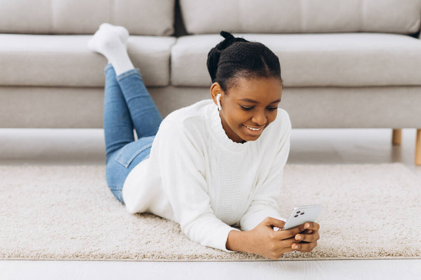 Sorridente ragazza nera messaggistica su smartphone a casa, rilassarsi sul comodo divano, godersi il fine settimana, spazio copia - Foto, immagini