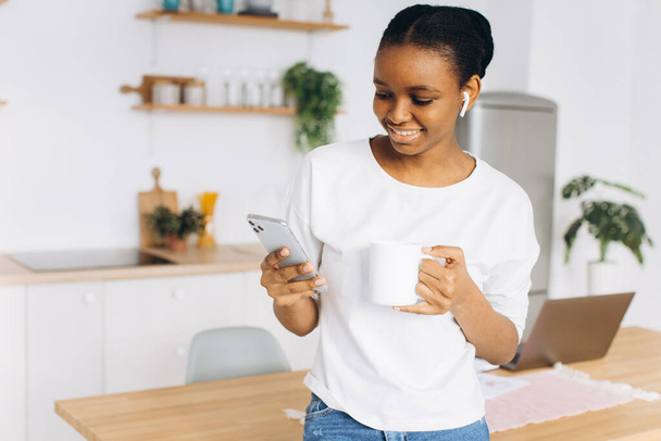 Portret van een vrolijke jonge zwarte vrouw met een telefoon en het drinken van koffie in de keuken. - Foto, afbeelding