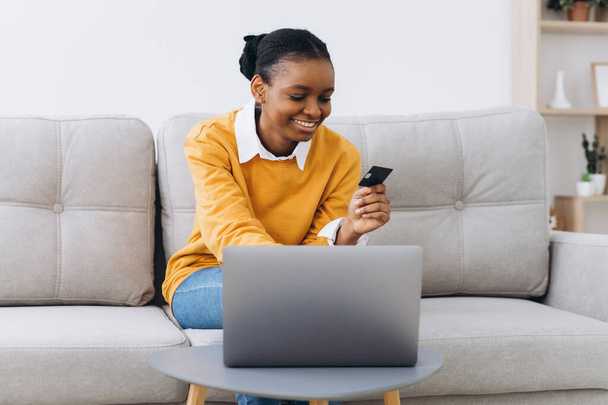 Compras en línea. Chica negra joven positiva usando el ordenador portátil y la tarjeta de crédito en casa, haciendo compras en Internet, sentado en el sofá en la sala de estar
 - Foto, imagen