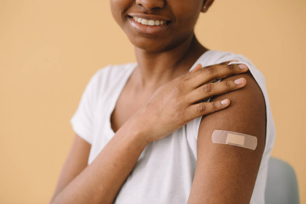 Молодая афроамериканка радуется первой вакцинации, носит пластырь, поднимает рукав и рассказывает друзьям об инъекции. Изолированный на бежевом фоне. - Фото, изображение