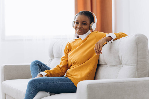 Rest And Relax Concept. Calm black woman sitting on bean bag, listening to music, audio book, podcast, enjoying meditation for sleep and peaceful mind in wireless headphones, leaning back, copy space - Photo, Image