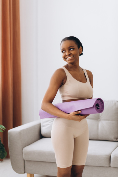 Inländisches Training. Junge sportliche schwarze Frau hält zu Hause eine Yogamatte in der Hand, trainiert im Wohnzimmer - Foto, Bild