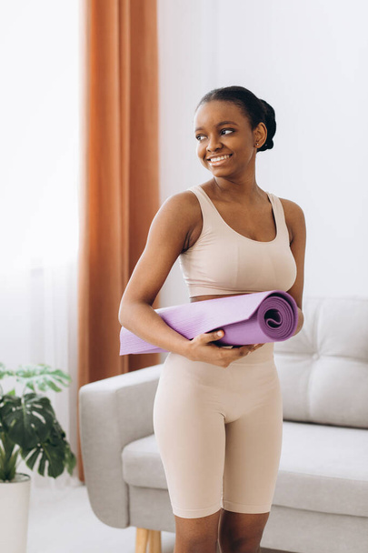 Domácí cvičení. Mladý sportovní černošky žena drží jóga podložka doma, trénink v obývacím pokoji - Fotografie, Obrázek