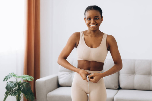 Jovem mulher negra mede a cintura no fundo da sala de estar. O conceito de dieta e perda de peso. - Foto, Imagem