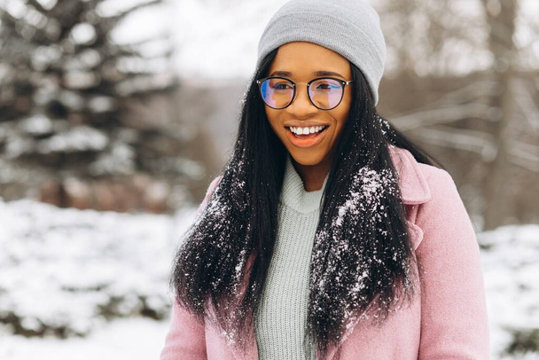 Portrét šťastné pozitivní dívky, africké etnické Afroamerické mladé ženy se sněhem na vlasech a v rukavicích se usmívá v zimním zasněženém parku ve sněhu za chladného mrazivého dne v teplých šatech. Zimní koncept - Fotografie, Obrázek