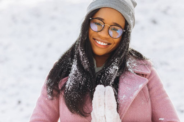 Ritratto di felice ragazza positiva, africana di etnia afro-americana giovane donna con la neve sui capelli e nei guanti sorride al parco innevato invernale nella neve nella fredda giornata gelida in abiti caldi. Idea invernale - Foto, immagini