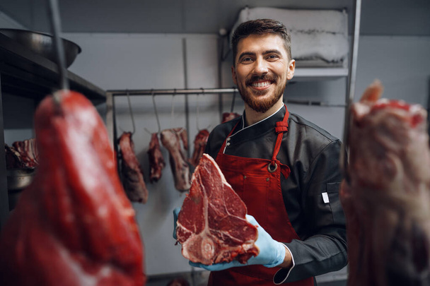 Young butcher holding raw meat steaks in fridge of grocery shop - Photo, image