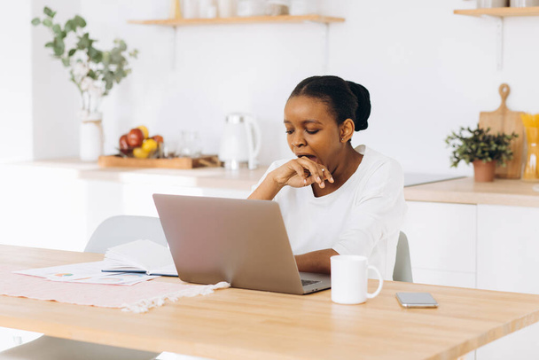 Une jeune femme noire est assise dans la cuisine à travailler sur un ordinateur portable, elle est fatiguée, bâille et veut dormir. - Photo, image