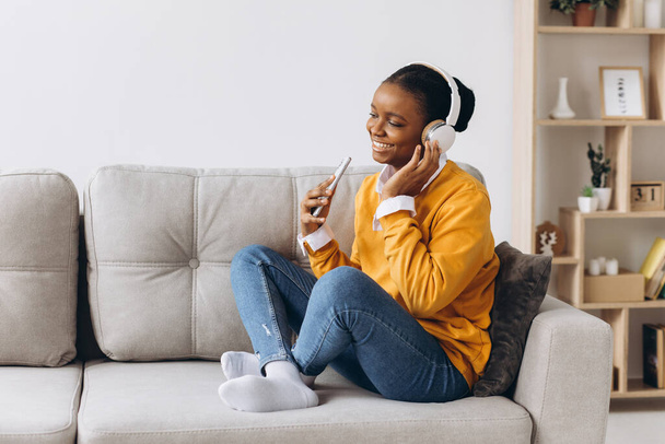 People, technology and leisure concept - happy african american young woman sitting on sofa with smartphone and headphones listening to music at home - Fotografie, Obrázek