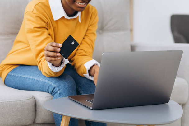 Online winkelen. Positief jong zwart meisje met behulp van laptop en creditcard thuis, het maken van aankopen in het internet, Zittend op de bank in de woonkamer - Foto, afbeelding