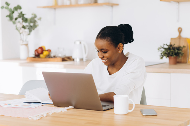 Fiatal fekete nő ül a konyhában, és dolgozik laptop otthonról. - Fotó, kép