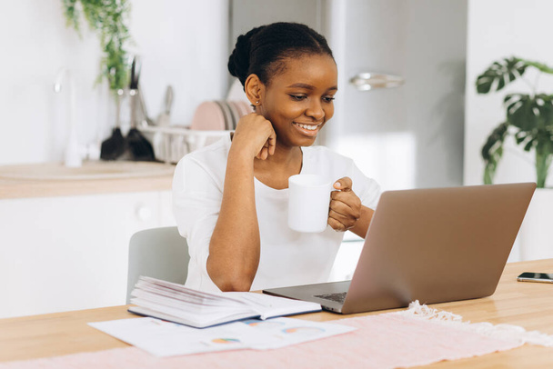 Jeune femme noire assise dans la cuisine buvant du café et parlant sur appel vidéo sur ordinateur portable. - Photo, image