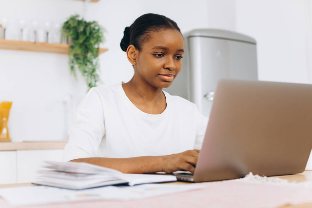 Genç siyahi bir kadının portresi mutfakta oturuyor ve evden bilgisayarla çalışıyor..  - Fotoğraf, Görsel