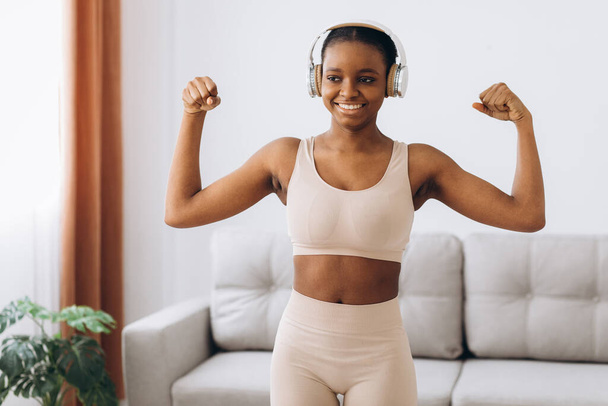 ヘッドフォンの若い黒人女性は音楽に耳を傾け、リビングルームで自宅でワークアウトを行います。ホームトレーニング. - 写真・画像