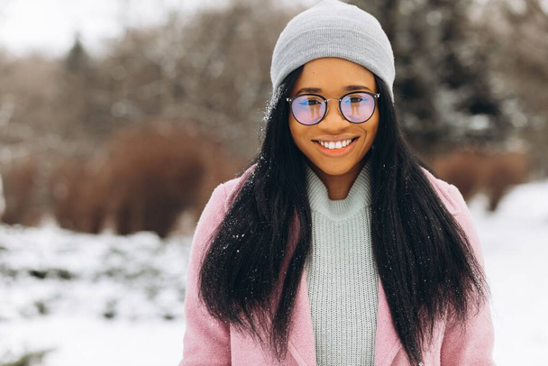 Portrét šťastné pozitivní dívky, africké etnické Afroamerické mladé ženy se sněhem na vlasech a v rukavicích se usmívá v zimním zasněženém parku ve sněhu za chladného mrazivého dne v teplých šatech. Zimní koncept - Fotografie, Obrázek
