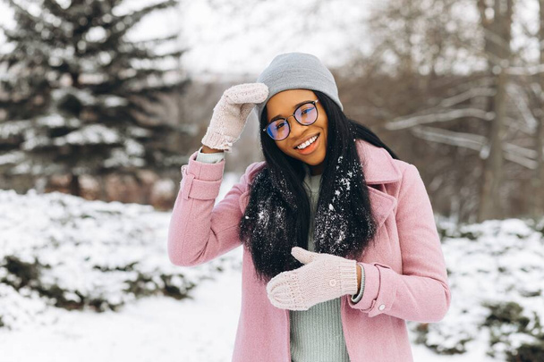 Portret van gelukkig positief meisje, Afrikaanse etnische Afro-Amerikaanse jonge vrouw met sneeuw op haar haar en in handschoenen glimlacht in de winter besneeuwd park in de sneeuw op koude ijzige dag in warme kleren. Winter concept - Foto, afbeelding