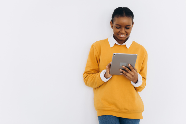 Happy black student girl in a yellow jacket holds a tablet and shows cheers on a white background - Foto, imagen