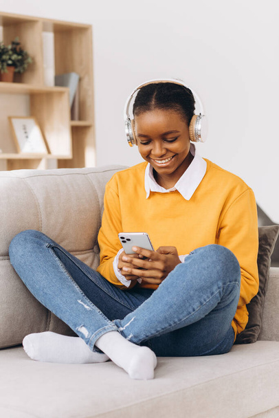 ihmiset, teknologia ja vapaa-ajan konsepti onnellinen afrikkalainen amerikkalainen nuori nainen istuu sohvalla älypuhelimen ja kuulokkeiden kuunnellen musiikkia kotona
 - Valokuva, kuva