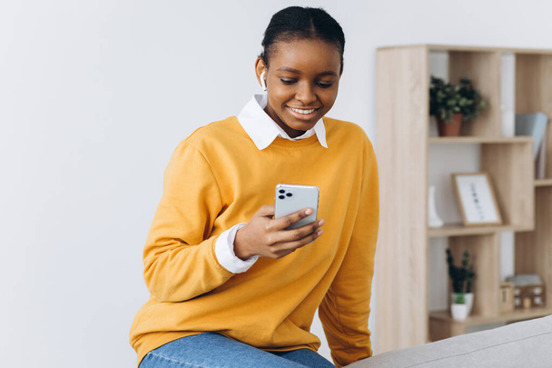 Sourire fille noire messagerie sur smartphone à la maison, détente sur un canapé confortable, profiter du week-end, copier l'espace - Photo, image